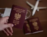 Dłoń trzyma polski paszport. W tle maseczka, samolot i inne paszporty.