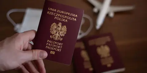 Dłoń trzyma polski paszport. W tle maseczka, samolot i inne paszporty.