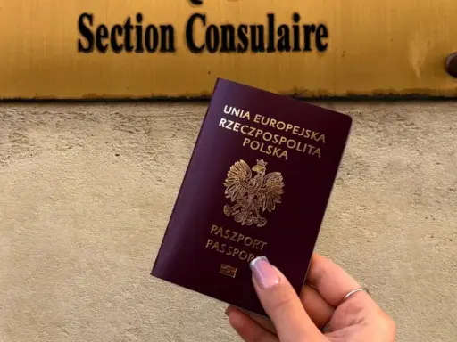 Dłoń trzyma polski paszport na tle tablicy z napisem "Section Consulaire".
