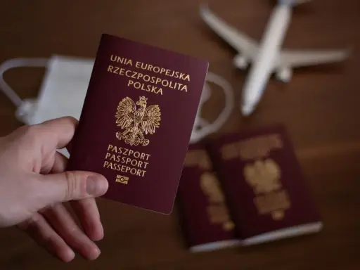 Dłoń trzyma polski paszport. W tle maseczka, samolot i inne paszporty.