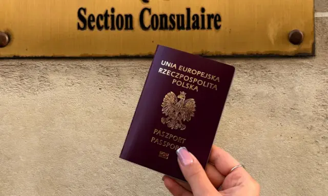 Dłoń trzyma polski paszport na tle tablicy z napisem "Section Consulaire".