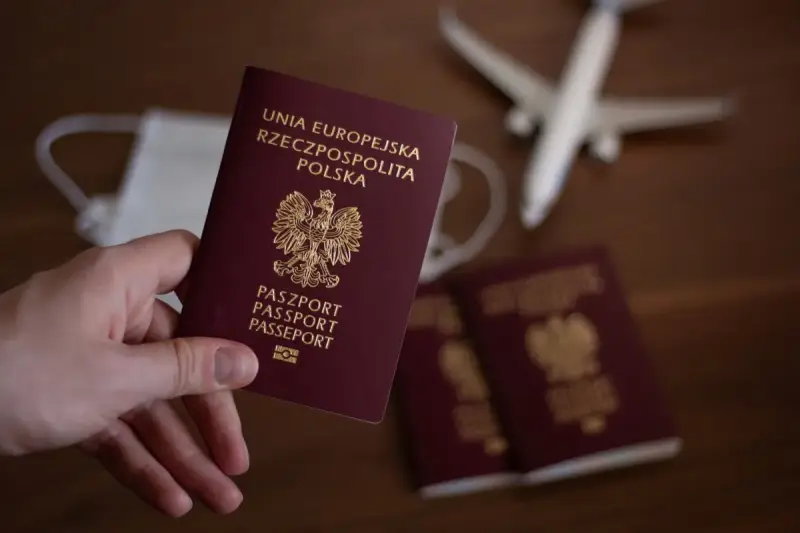 Dłoń trzyma polski paszport. W tle maseczka, samolot i inne paszporty.